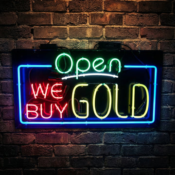 Neon sign on a brick wall that reads 'Open We Buy Gold'.